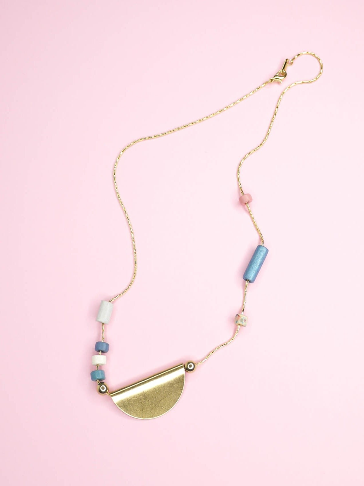 Lightweight & delicate ceramic beaded necklace with a brass semi circle pendant in the centre with ceramic tube beads on either side in pink, blues, white & silver, strung on a thin gold plated snake chain.