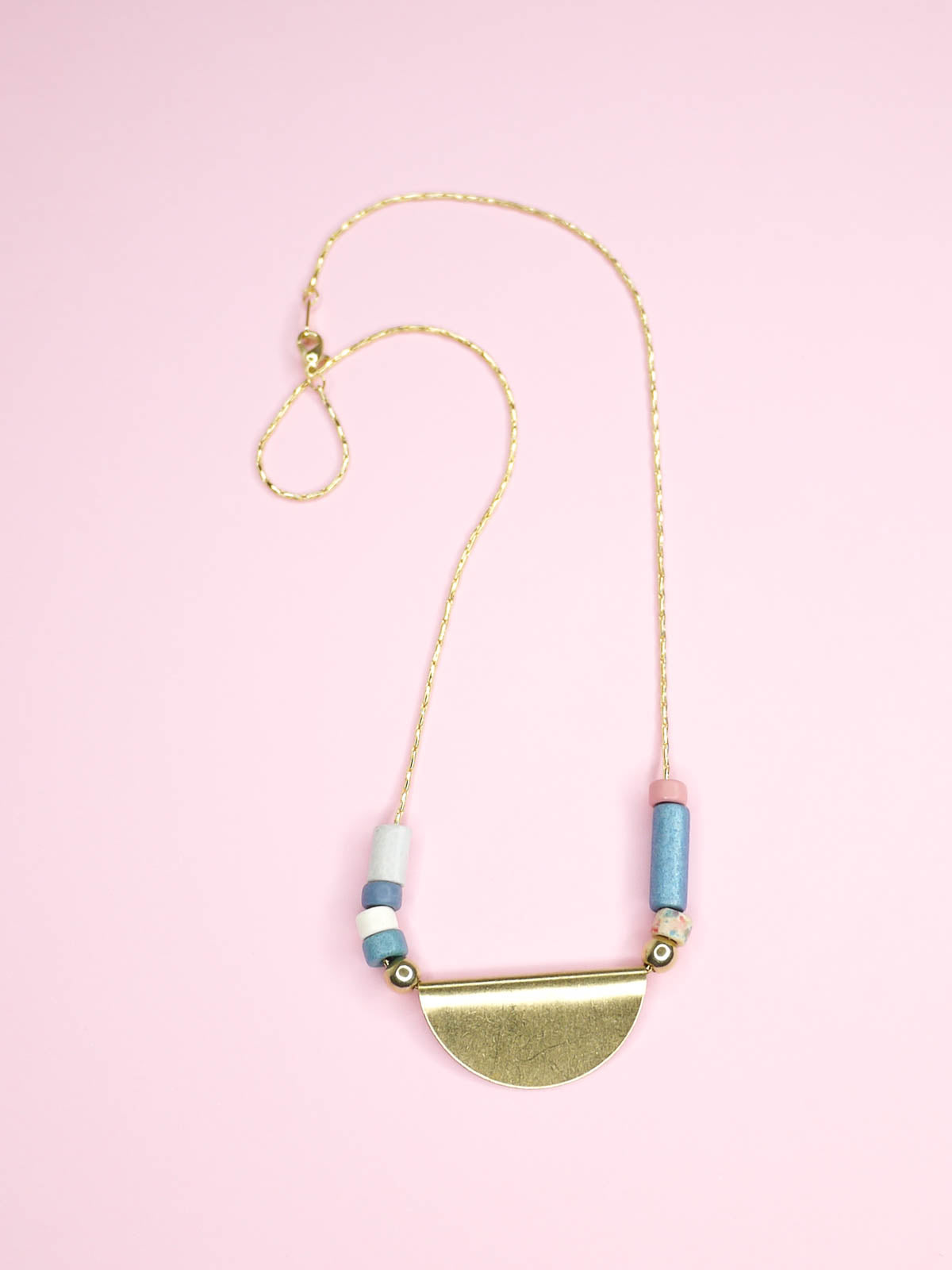 Lightweight & delicate ceramic beaded necklace with a brass semi circle pendant in the centre with ceramic tube beads on either side in pink, blues, white & silver, strung on a thin gold plated snake chain.