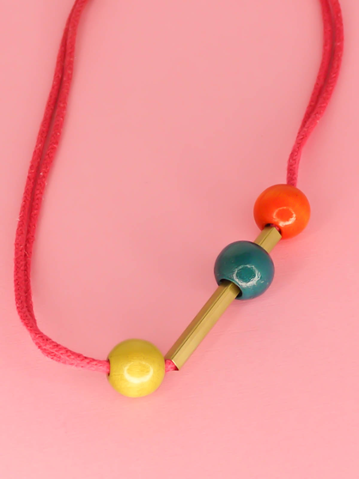 Geometric statement necklace in hot pink cotton rope with a brass bar in the centre with contrasting wooden beads on either side in orange and green and a turquoise bead over the brass bar with a brass clasp