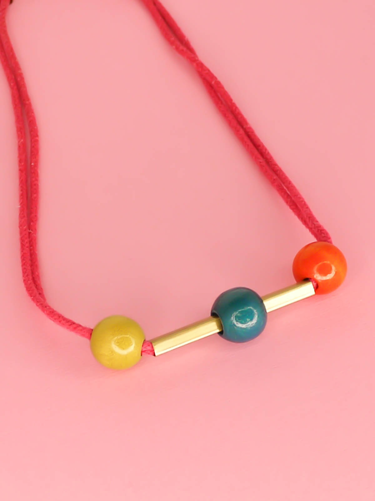 Geometric statement necklace in hot pink cotton rope with a brass bar in the centre with contrasting wooden beads on either side in orange and green and a turquoise bead over the brass bar with a brass clasp