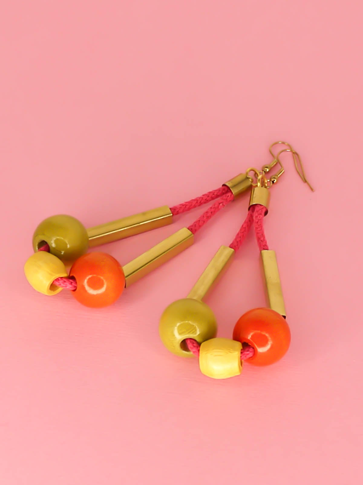 Long bead dangle statement earrings with wooden beads in green, orange & yellow & brass tube beads strung on hot pink cotton rope with brass end caps, hung from gold plated brass earring hooks.