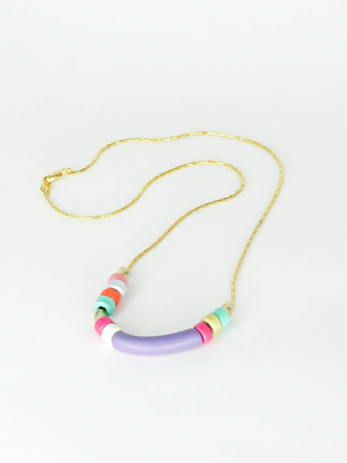 Dainty beaded ceramic necklace with a lilac curved bar bead, surrounded by textured brass tube beads and colourful ceramic beads in pink, turquoise, orange & white. Hung from a gold plated snake chain.