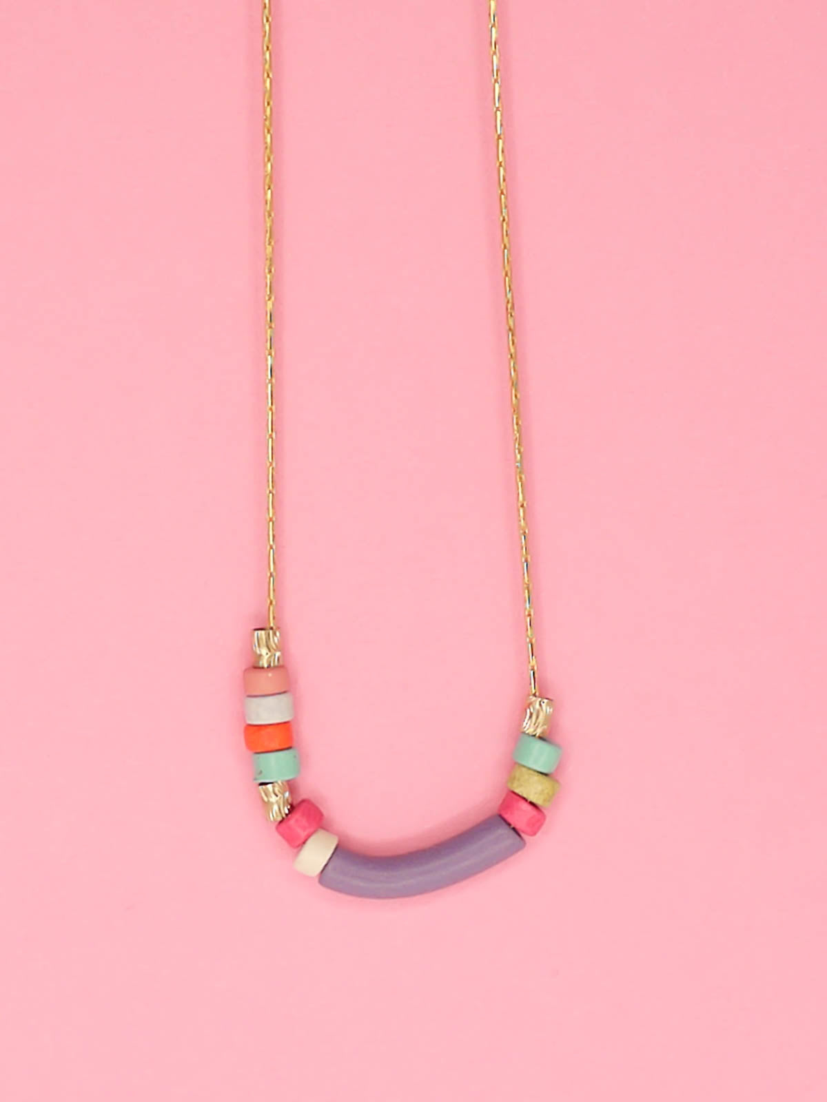 Dainty beaded ceramic necklace with a lilac curved bar bead, surrounded by textured brass tube beads and colourful ceramic beads in pink, turquoise, orange & white. Hung from a gold plated snake chain.