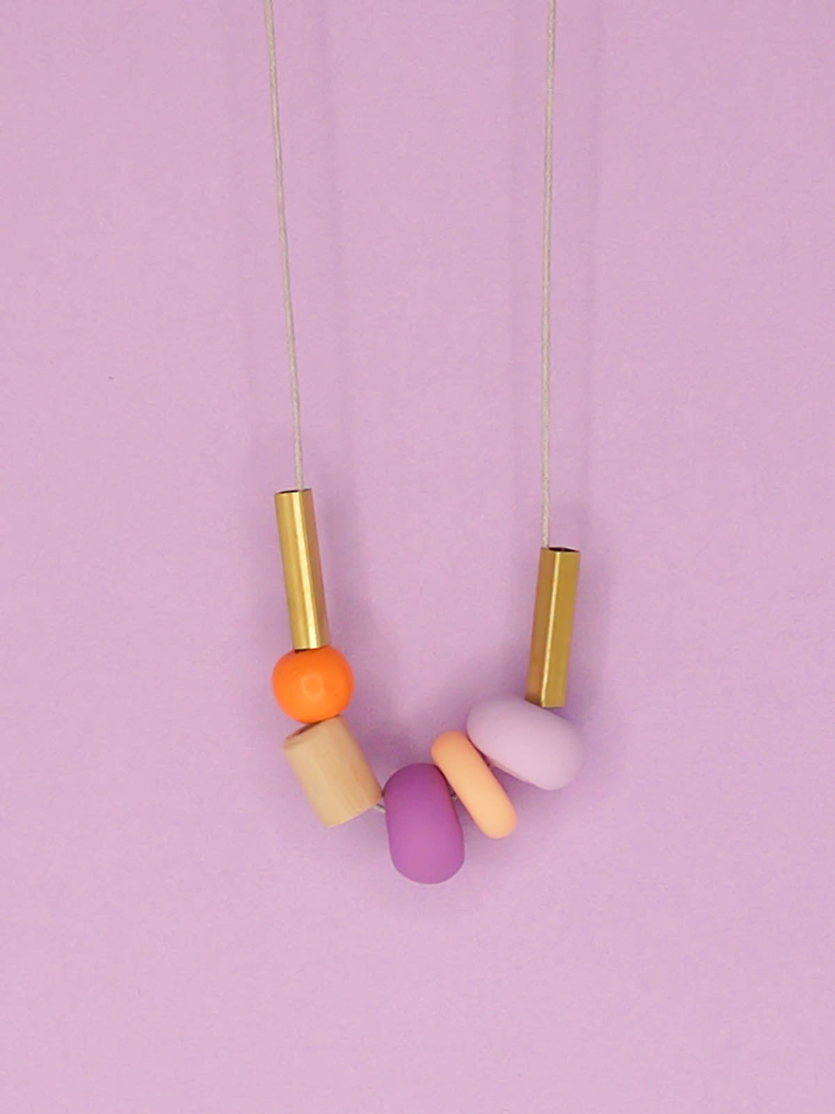 Lightweight geometric colourful bead necklace with 3 handmade polymer clay beads in peach, lilac & purple- wood tube bead & orange wood bead, brass tube beads on either side, strung on a cotton cord with a wood bead knotted at the end  