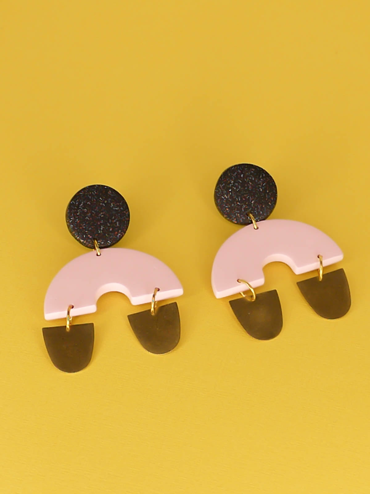 Large geometric arch dangle chandelier earrings with a black glitter stud, connected to a pink semi circle with two brass semi circles hanging off either side of the arch. Made from polymer clay with stainless steel posts.